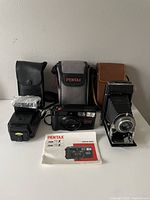 Photo showing Pentax 35mm camera with padded case, Kodak folding camera in leather case, Canon flash, and Pentax manual laid out on surface.