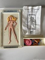 Photo of vintage plastic flip and fold fashion doll with blonde hair on original white display tray, instruction sheet, and small box of accessories