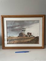 Frontal view of the framed watercolour print depicting grain elevators under a cloudy sky, placed on a flat surface next to a pen for scale.