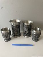 Set of 4 German pewter cups showing two sizes with detailed relief artwork, placed against a gray wall with a pen for size reference.