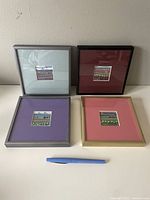 Four framed 6x6 inch gouache paintings with colorful mats and silver or gold frames on white surface with blue pen for scale