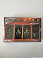 Front of sealed LEGO Jurassic Park mini figures limited edition set showing 3 character minifigures and dinosaur figures through window on red/black packaging.