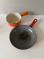 Two small Le Creuset cookware items: a red frying pan with grey interior and an orange saucepan with white interior.