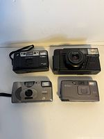 Front view of four 35mm point and shoot film cameras sitting on a white surface labeled Canon, Pentax, Kodak, Nikon.