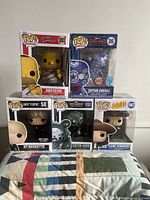 Five Funko Pop figures boxed and displayed: Homer Simpson Obeseus, Captain America translucent blue, Hot Topic Nerdette, Doctor Doom, Elaine from Seinfeld.