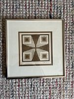 Framed abstract print of geometric art in neutral tones on textured carpet background.
