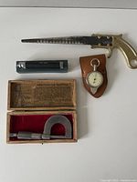 Top view of lot including a serrated metal hand tool with brass pistol grip handle, leather case stop watch, small black cylindrical tool, and boxed micrometer.