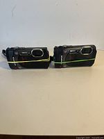 Two Olympus Stylus TG-870 cameras side by side, front lens and flash area visible with rubber bands securing the battery door.