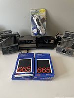 Five vintage Polaroid cameras on a table including box style and Land models, with four Polaroid 665 film packs and a white device in packaging.
