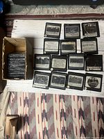 Display of 15 individual antique glass slides laid out on a wooden surface with a cardboard box containing additional slides.