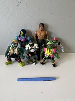 Front view of all seven figures showing their relative size with blue pen for scale, standing on white surface against gray background.