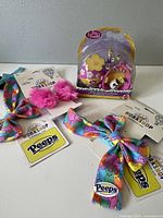 Photo showing Build-A-Bear Workshop accessories: three colorful patterned bows with rainbow Peep design, two pink fuzzy hair ties, all with Peeps branding visible.