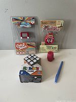 Overview photo showing the entire toy lot with all items including World's Smallest Etch A Sketch, Rubik's Cube, Mini Barrel of Monkeys, The Office talking keychain, and a blue pen-like object.