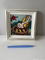 Framed vintage cross-stitch artwork showing a bird feeding chicks in a nest, with colorful stitching and a white wooden frame.