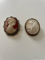 Front view of two vintage cameo brooches, one with a female profile and one with a scene of a woman near a house