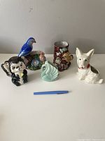Six vintage ceramic planters and vases arranged on a white surface, various animal-shaped and floral designs visible, including blue bird, dog, skunk, swan, brown floral vase, and floral planter with bird figurines.