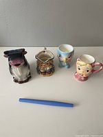 Four vintage Japanese ceramic items displayed in a row on a white surface with a blue pen for scale, showing the cow creamer, Toby jug, bluebird egg cup, and Miss Cutie Pie mug in full view.