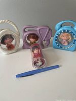 Front view of the four Little Kiddles dolls each in unique plastic cases shaped like a basket, elephant, telephone, and a clear cylinder, small blue pen shown for scale.