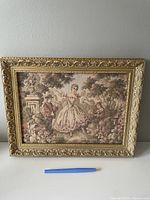 Photo of a vintage French tapestry in a gilded ornate frame, placed against a neutral wall with a pen for scale.