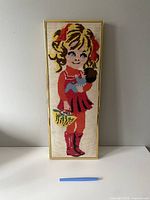 Full view of framed needlepoint showing girl in red dress holding doll and basket, with pen for scale.