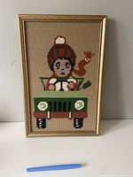 Framed needlepoint artwork showing a child in a green car against a beige fabric background.