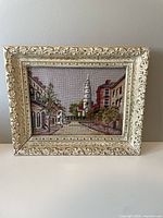 Front view of vintage needlepoint artwork in ornate cream carved frame showing street and church steeple scene.