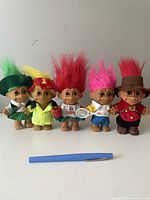 Five Russ troll dolls in colorful costumes and different hair colors arranged in a row against a white wall, pen laid in front for size reference