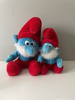 Front view of two vintage Papa Smurf plush toys with red hats, white beards, and blue bodies in different sizes.