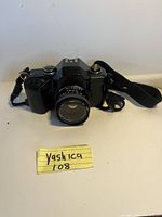 Front view of the black Yashica 108 35mm SLR camera with attached lens and strap, placed on a white surface with a handwritten label showing 'Yashica 108'.