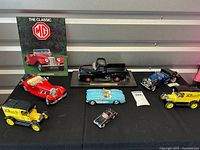 Full collection of 7 die cast model cars displayed on black cloth with metal panel background, includes Chevrolet Corvette, Ford Model T Van, Mercedes Roadster, Duesenberg, and Ford Pickup truck