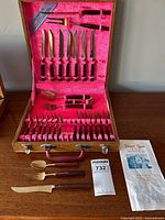 Top view of open wooden case displaying rosewood-handled steak knives, forks, serving utensils and a metal label inside the lid.