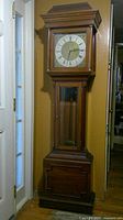 Full view of tall wooden grandfather clock standing against wall near door. Clock has glass front panel and wooden case.