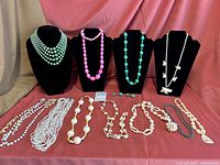 Full view of 13 costume jewelry necklaces displayed on black bust forms and laid flat.