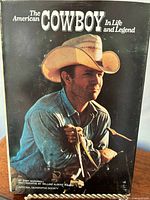 Book titled 'The American Cowboy In Life and Legend' showing cover photo of a man in a cowboy hat holding reins of a horse or cattle.
