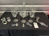 Full set of etched glass decanter with stopper, six matching etched glasses, glass owl figurine, three additional decanter tops laid out on black cloth