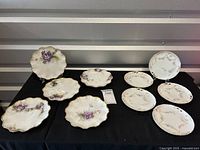 Overview photo showing 9 plates arranged on a black cloth, displaying two different floral patterns, all with scalloped gold edges.