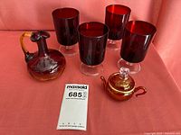 Four maroon goblets each with a clear pedestal base, a small maroon glass decanter with handle and stopper, and a maroon sugar bowl with handle and lid, all displayed on a pink fabric background.