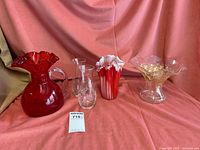 Lot of five glass items: red fluted pitcher, two clear etched glass vases, clear gold etched vase, red and white fluted blown vase on pink cloth background