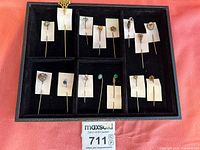 Full tray of stickpins arranged in 15 compartments on black velvet lining with white paper holders.