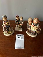 Three vintage Hummel ceramic figurines depicting children in traditional scenes: feeding chickens, playing soldiers, and going to market with a basket. All in mint condition, displayed on a wooden surface with a Maxsold lot tag visible.