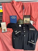 Full lot display with uniform jacket, helmet, books, flag, medal case and mug on red cloth backdrop