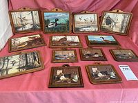 Full view of all 13 wood plaques arranged on pink fabric showing various waterfowl in painted scenes, including large and small plaques, plus the book not visible in this image.