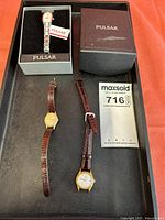 Overview of three women's watches: one Pulsar in box with broken band, two Seiko with brown leather straps on black tray with documentation