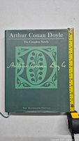 Front cover of the hardcover book showing title, decorative emblem, and Arthur Conan Doyle's signature with measuring tape for size reference.