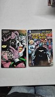 Two Batman hardcover comic books with visible covers. Left: Batman The Killing Joke Deluxe Edition featuring Joker holding a camera. Right: Batman Zero Year cover with Batman and other characters.