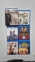 Top-down view of 6 Blu-ray cases laid flat: The Bourne Trilogy boxed set, two copies of The Bourne Identity and Supremacy, Oblivion, Iron Man, Cats & Dogs: The Revenge of Kitty Galore, John Carter.