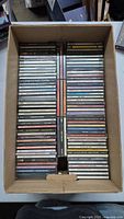 Full view of a box filled with four rows of assorted music CD jewel cases, arranged neatly side by side.