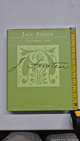 Front cover of the book showing the title 'Jane Austen The Complete Novels The Autograph Edition' with a green dust jacket and a decorative letter 'A'. A measuring tape indicates the size of the book.
