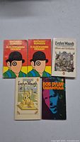 Five vintage paperback books arranged on a flat surface, including two copies of 'A Clockwork Orange' by Anthony Burgess, two books by Evelyn Waugh, and one by Bob Dylan.