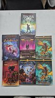 Seven Rick Riordan books laid out on a table with colorful mythological artwork on the covers.